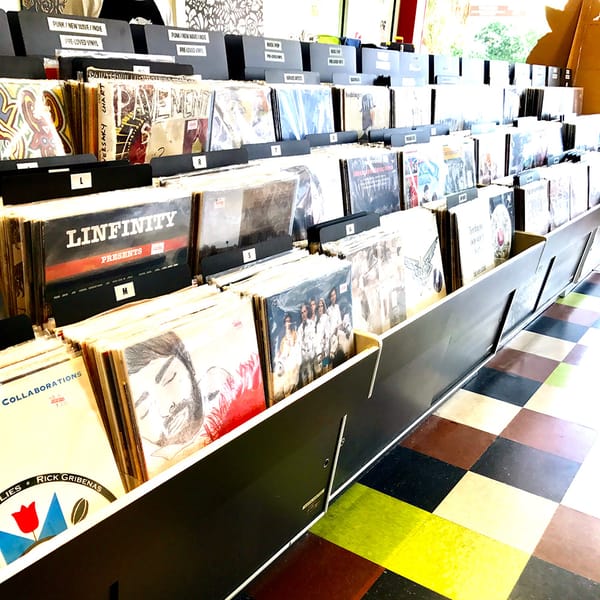 A photograph of a retail shelf full of records with a colorful checkerboard floor.